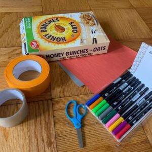 A box of tape, scissors, and markers are on a wooden floor.