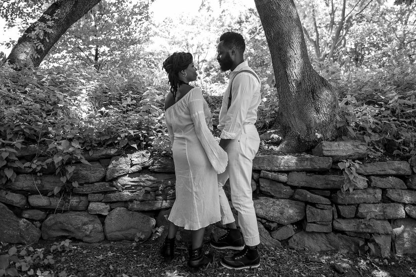 A black and white photo of a couple standing next to a stone wall.