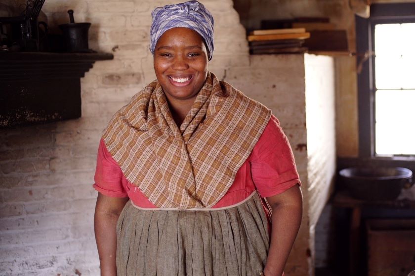 A woman in an old fashioned dress smiling in front of a fireplace.