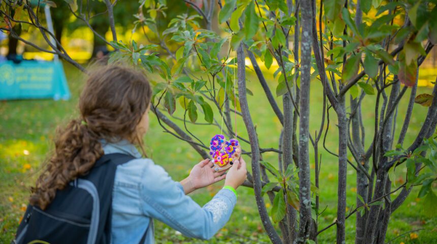 A girl with a backpack picking up a flower from a tree.