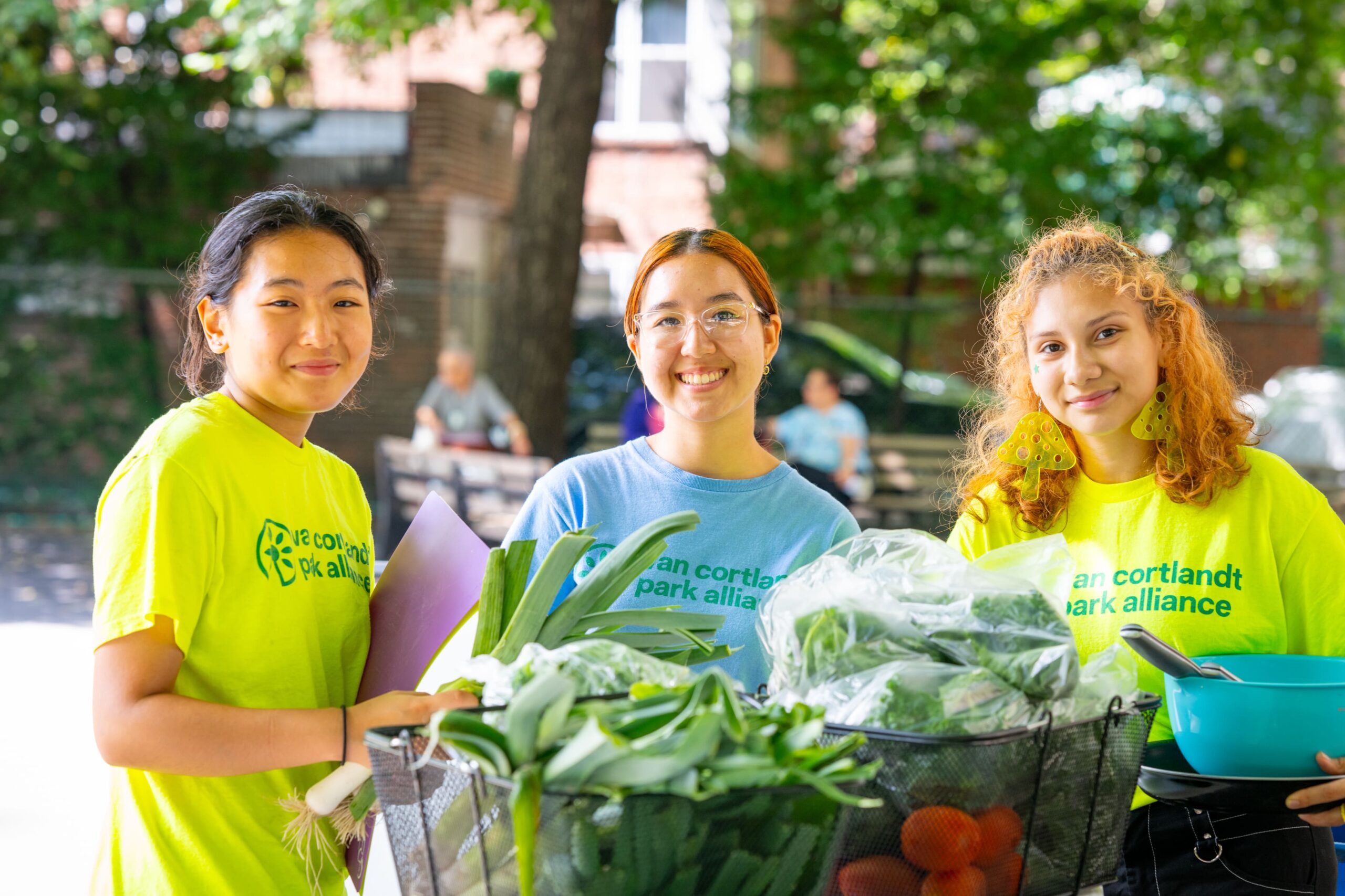 Three young women in yellow shirts standing next to a basket of vegetables.