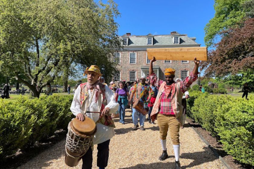 The Pinkster Players with a drummer in the lead, leading attendees on a procession in front of the Van Cortlandt House Museum.