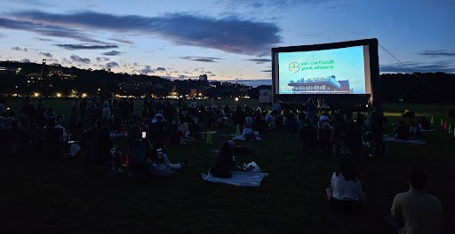 A movie screen with people sitting on a lawn in front of it.