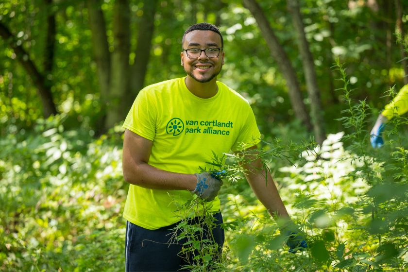 A young man removing weeds along a trail in the woods with a smile on his face.