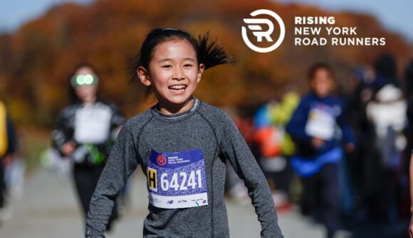 A young girl with a big smile on her face and a race bib on her sweatshirt running in the park.