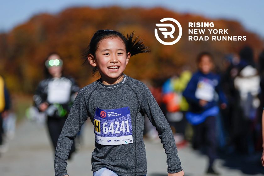 A young girl with a big smile on her face and a race bib on her sweatshirt running in the park.