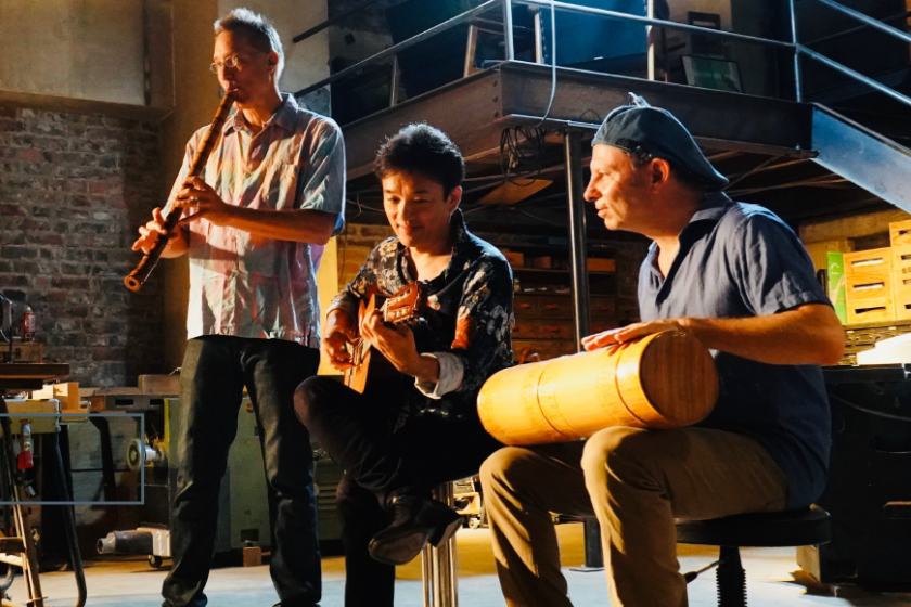 Three men playing instruments: shakuhachi flute, acoustic guitar, and Uduboo drum, an African Ud made from bamboo.