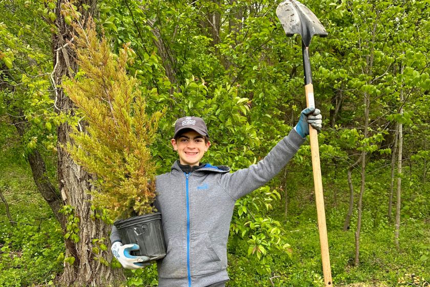 A young man holding a potted tree in one hand and holding up a shovel with the other hand with a big smile outside.