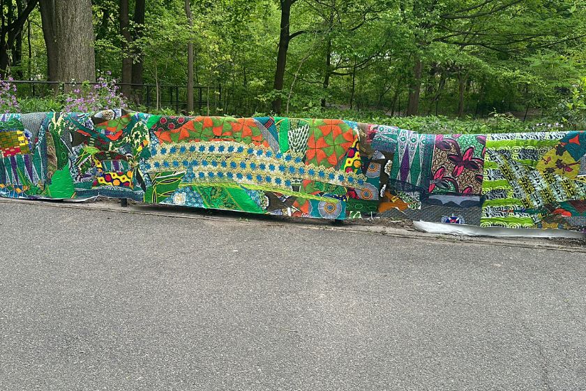 A painted tapestry over a fence in front of a paved path with woods behind