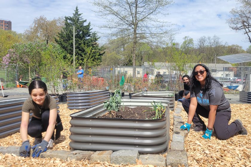 Two women on either side of a raised garden bed wearing gloves and smiling while working.