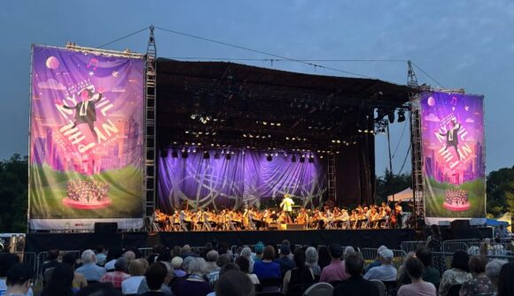 A large stage with purple banners on each side and a conductor in a white jacket with an orchestra at night.