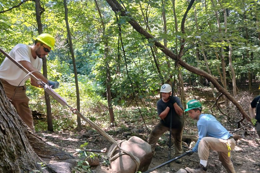 Three people in hardhats moving a boulder with tools and ropes.