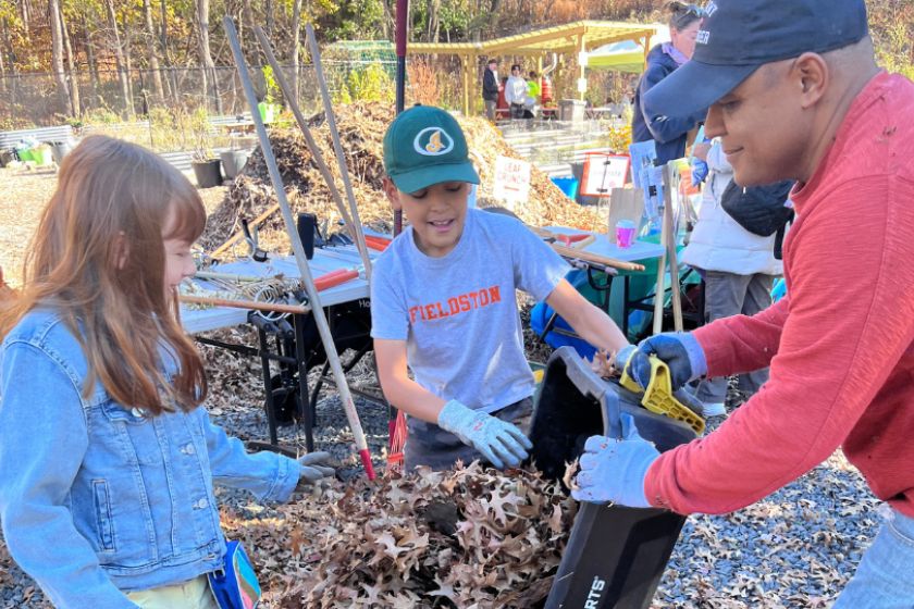 Two young children and a man outside around a bin with leaves in it.