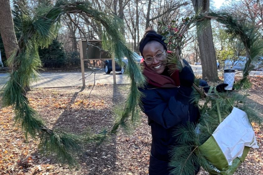 A woman in a black jacket and burgundy scarf smiling and holding a wreath while standing next to larger wreath.