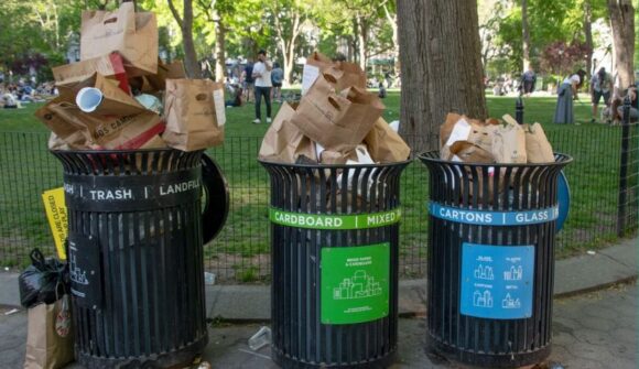 Three overflowing garbage cans along an asphalt path with grass and trees behind them.