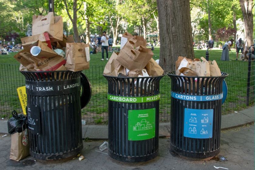 Three overflowing garbage cans along an asphalt path with grass and trees behind them.
