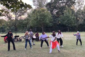 A man in a white outfit with a orange scarf around his waist teaching people to dance on the lawn