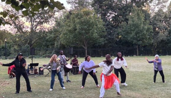 A man in a white outfit with a orange scarf around his waist teaching people to dance on the lawn