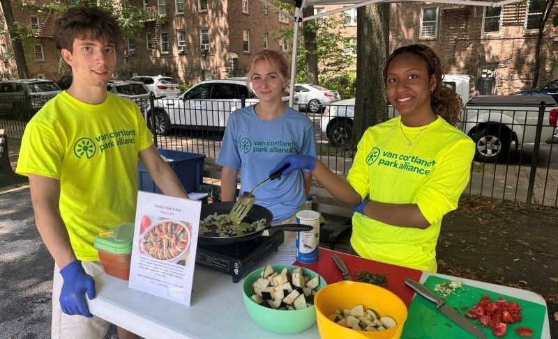 A young man in a yellow shirt with a young woman in a blue shirt and a young woman in yellow shirt with a table full of cut veggies with a pan cooking a meal.