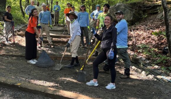 People standing on a inclined trail holding tools and smiling with trees in the background
